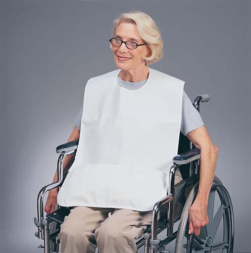 An older woman wearing glasses and a white bib sits in a wheelchair, facing forward against a plain background.
